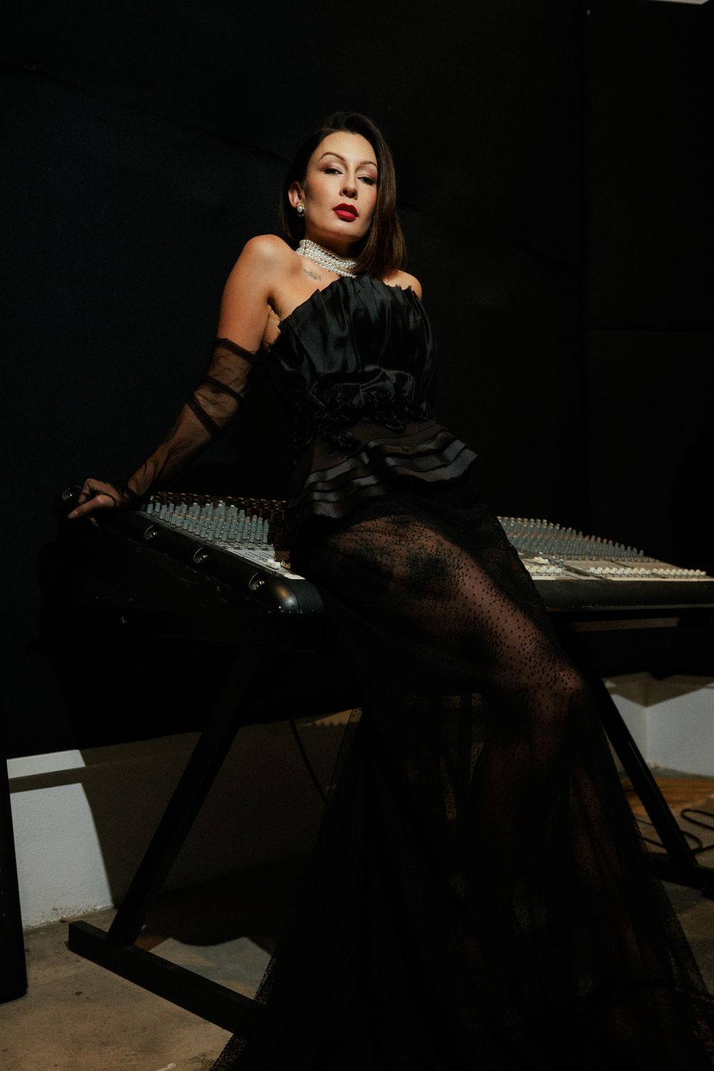 A person in a black evening gown and stockings poses dramatically on a grand piano in a dark moody setting.