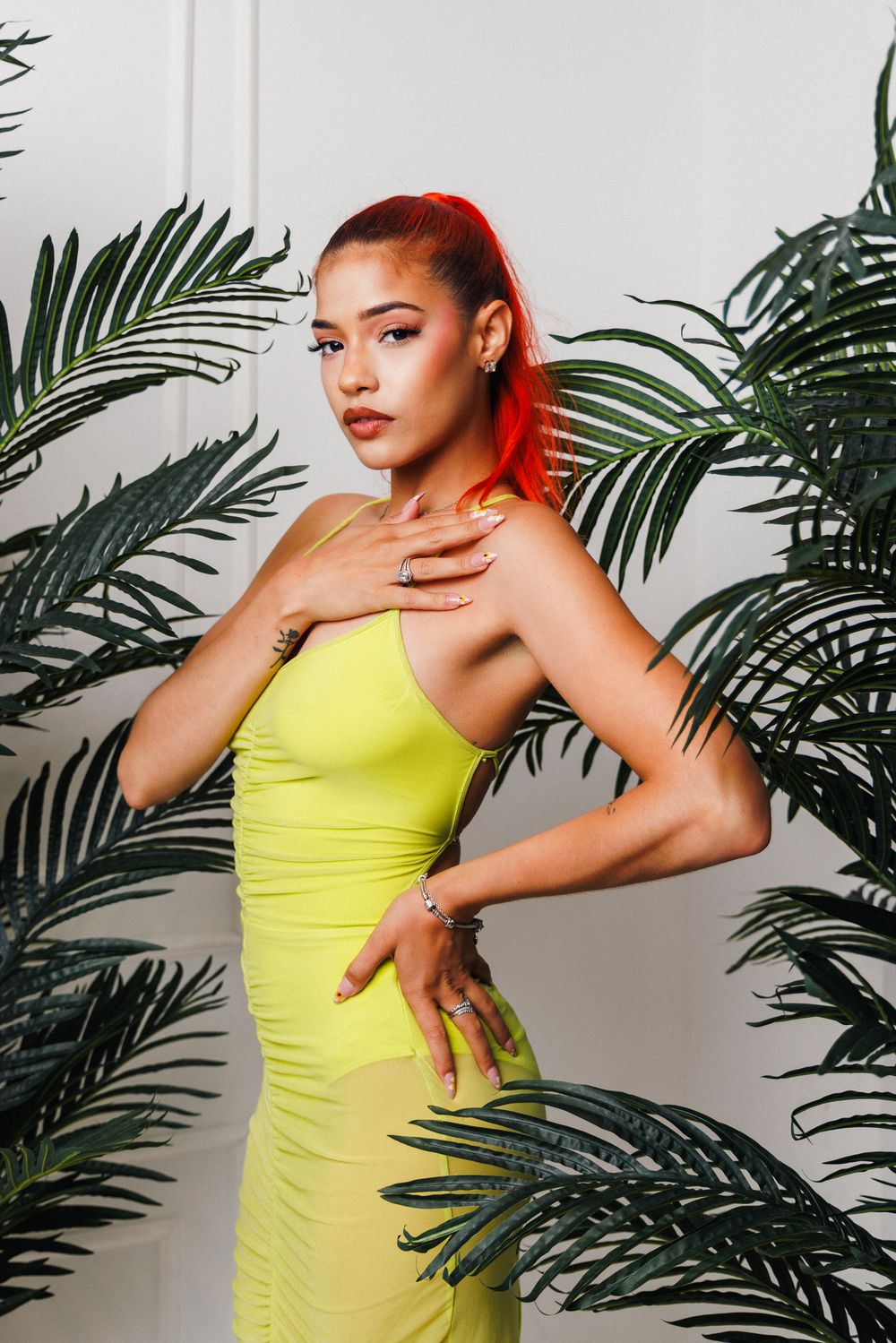Fashion model in bright yellow bodycon dress poses elegantly against tropical palm leaf backdrop.