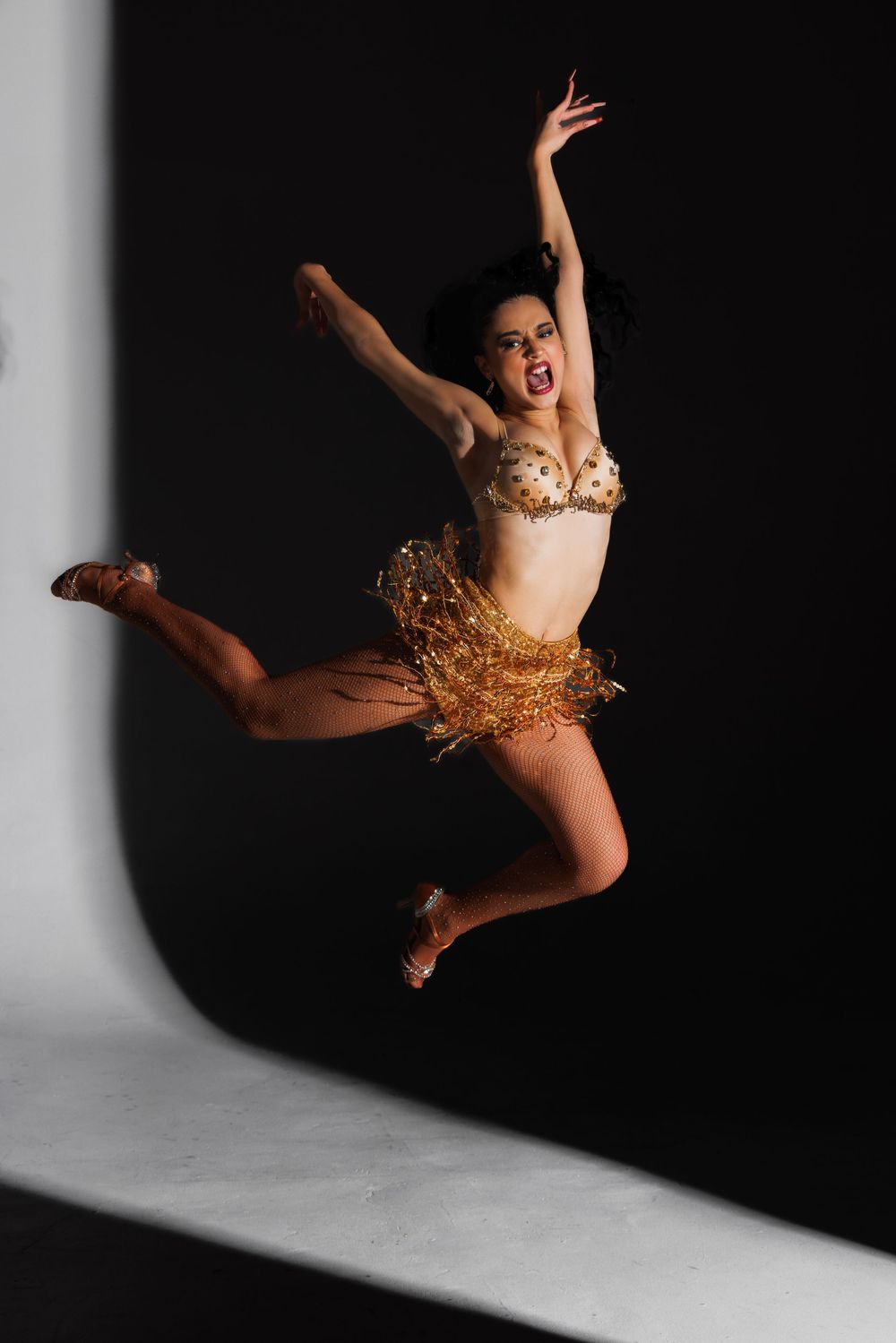 Energetic dancer in gold costume leaps gracefully through dark studio space in dramatic action sequence.