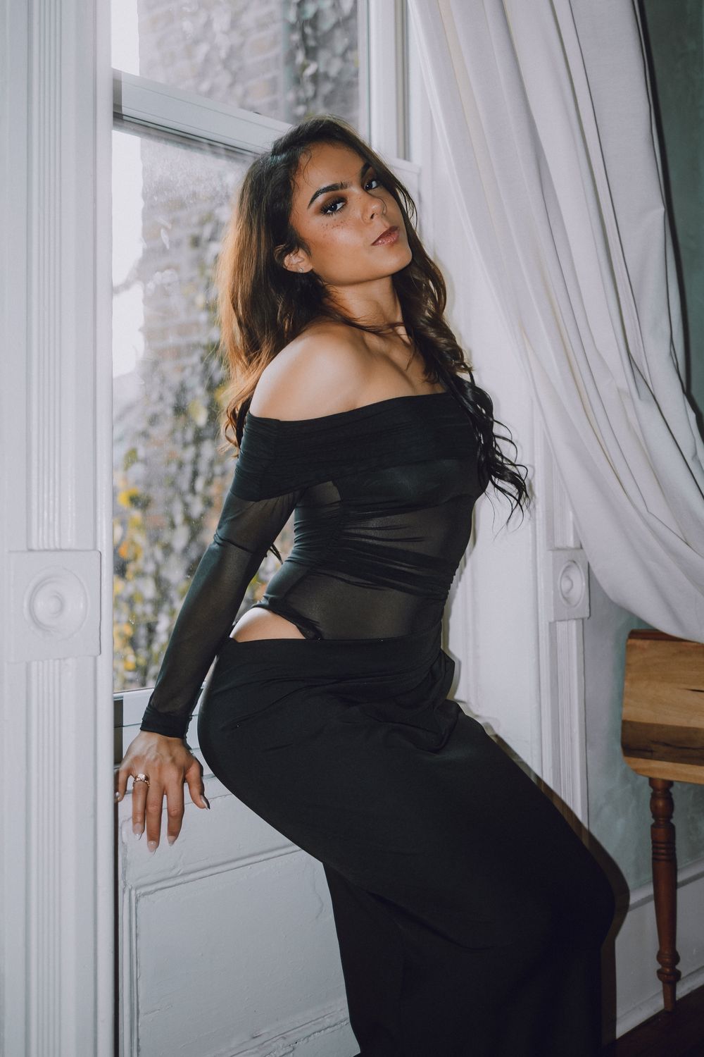 A woman in a black dress poses elegantly against white curtains in a vintage-style interior setting.