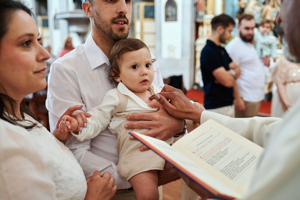 Parents holding baby in white christening outfit during Catholic baptism ceremony with priest reading from book.