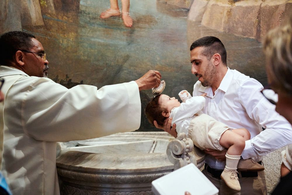 A priest performs a baptism, pouring holy water over an infant held by the father.