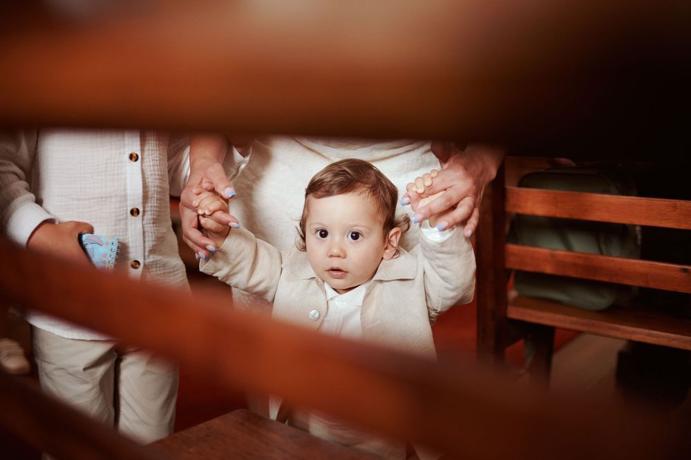 Curious baby in white baptism outfit standing between wooden pews, held by adults in a church.