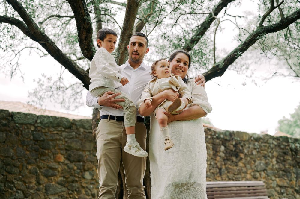 A family of four poses under a tree by a stone wall, parents each holding a young child.