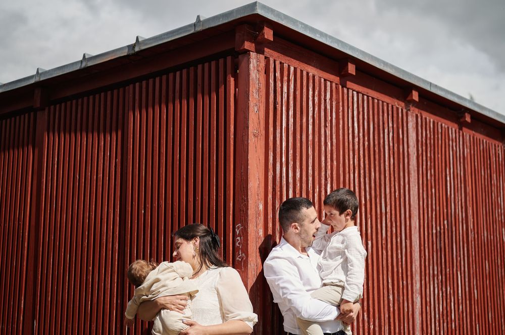 A family of four poses against a red corrugated metal wall, each parent holding a child.
