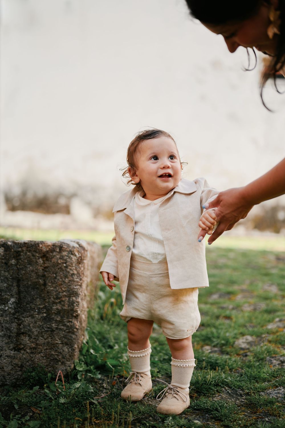 An adorable baby in a beige linen suit stands outdoors while being guided by an adult.
