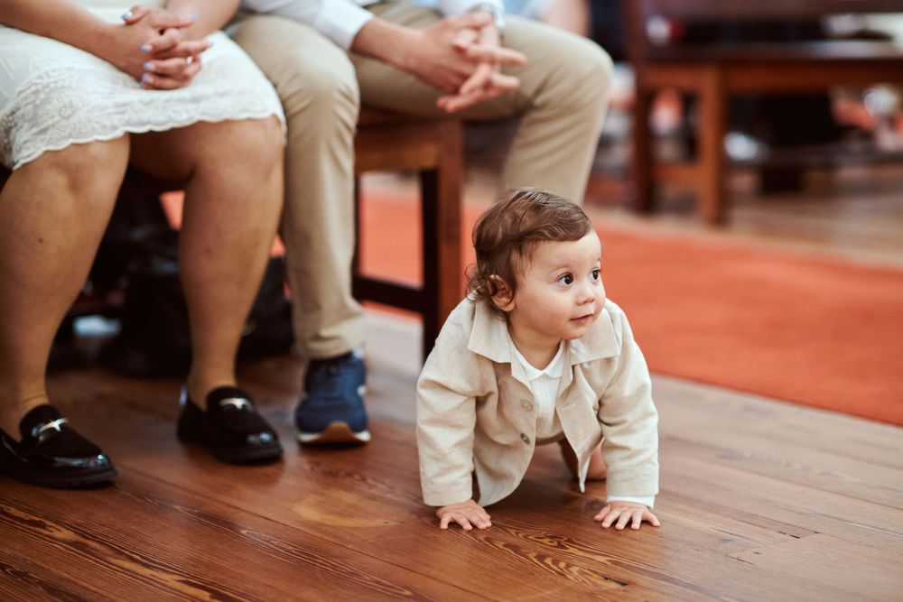 A curious baby in a beige outfit crawls on a hardwood floor during a baptism ceremony.