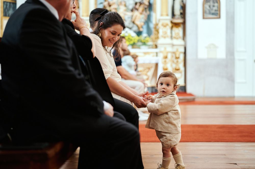 Smiling mother watching her toddler in a beige outfit standing in a church aisle during baptism.