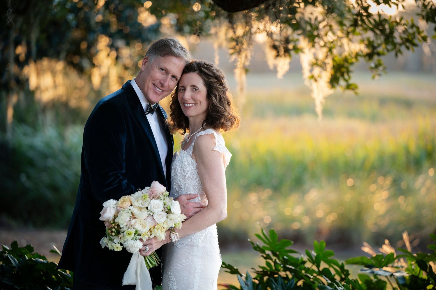 A newlywed couple poses outdoors at sunset with a white bouquet amid golden sunlight filtering through trees.