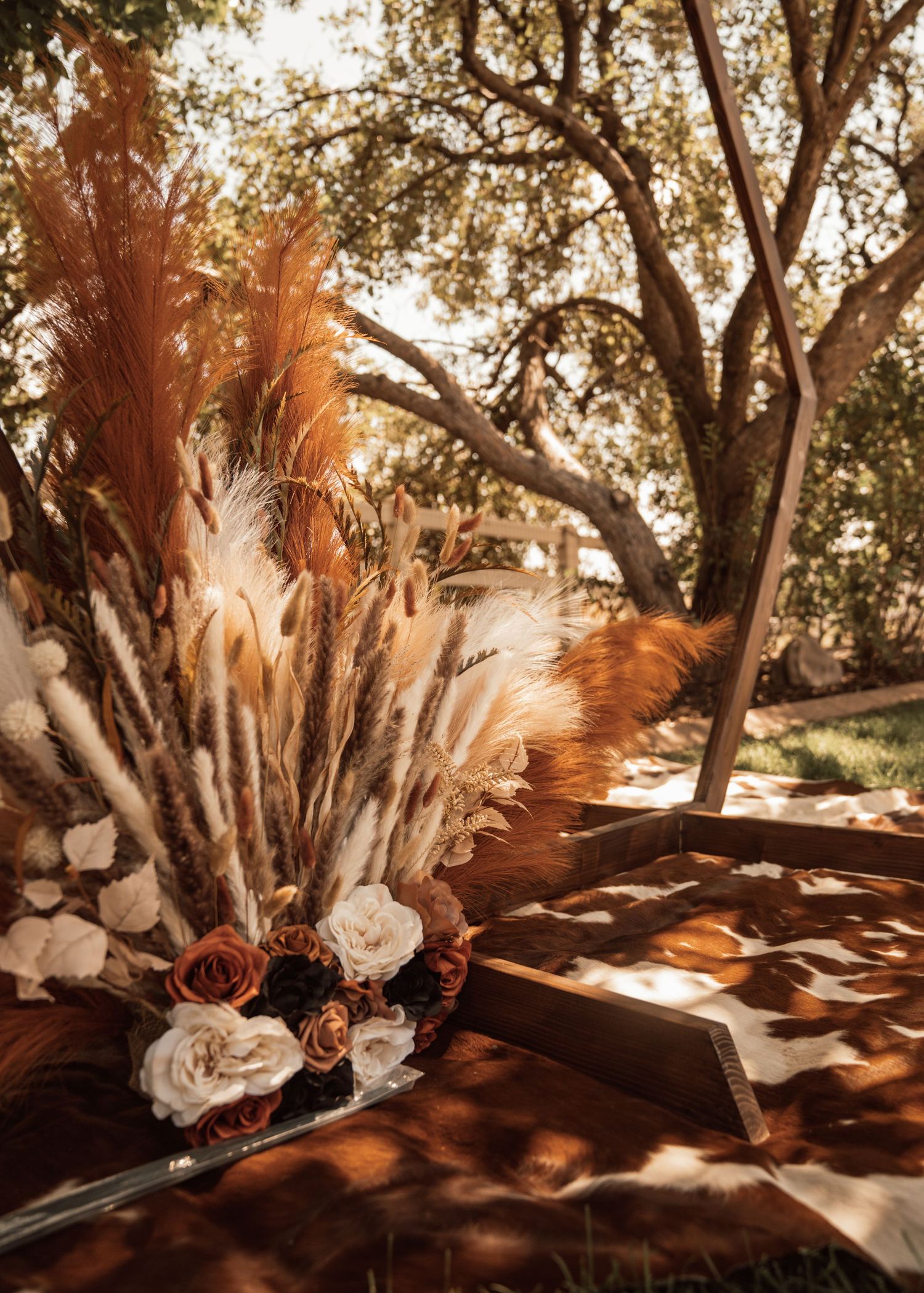Rustic wedding decor featuring dried pampas grass and white roses arranged on wooden surfaces in natural sunlight.