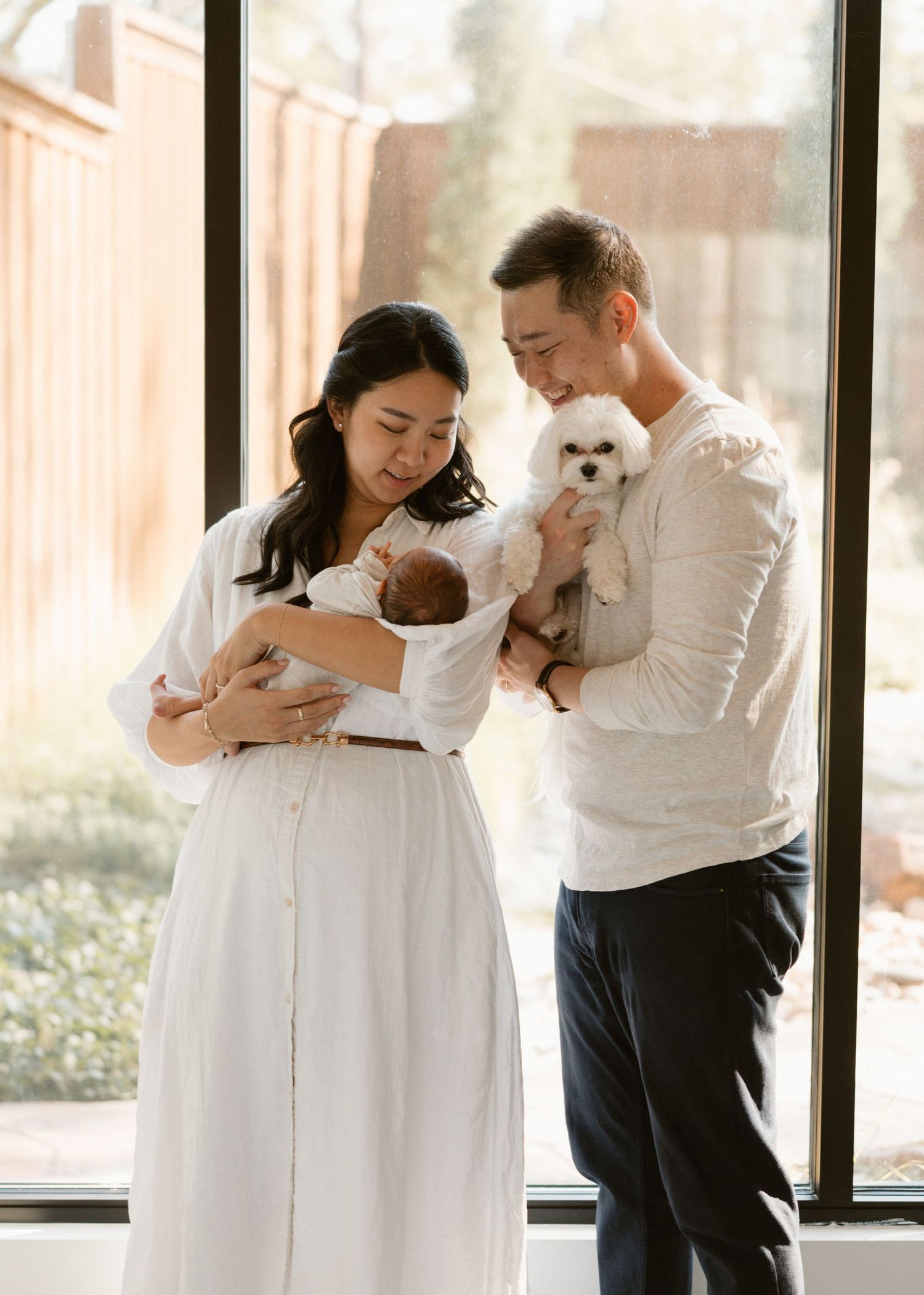 A couple in white and black attire embraces lovingly while holding a small white dog in a bright interior space.