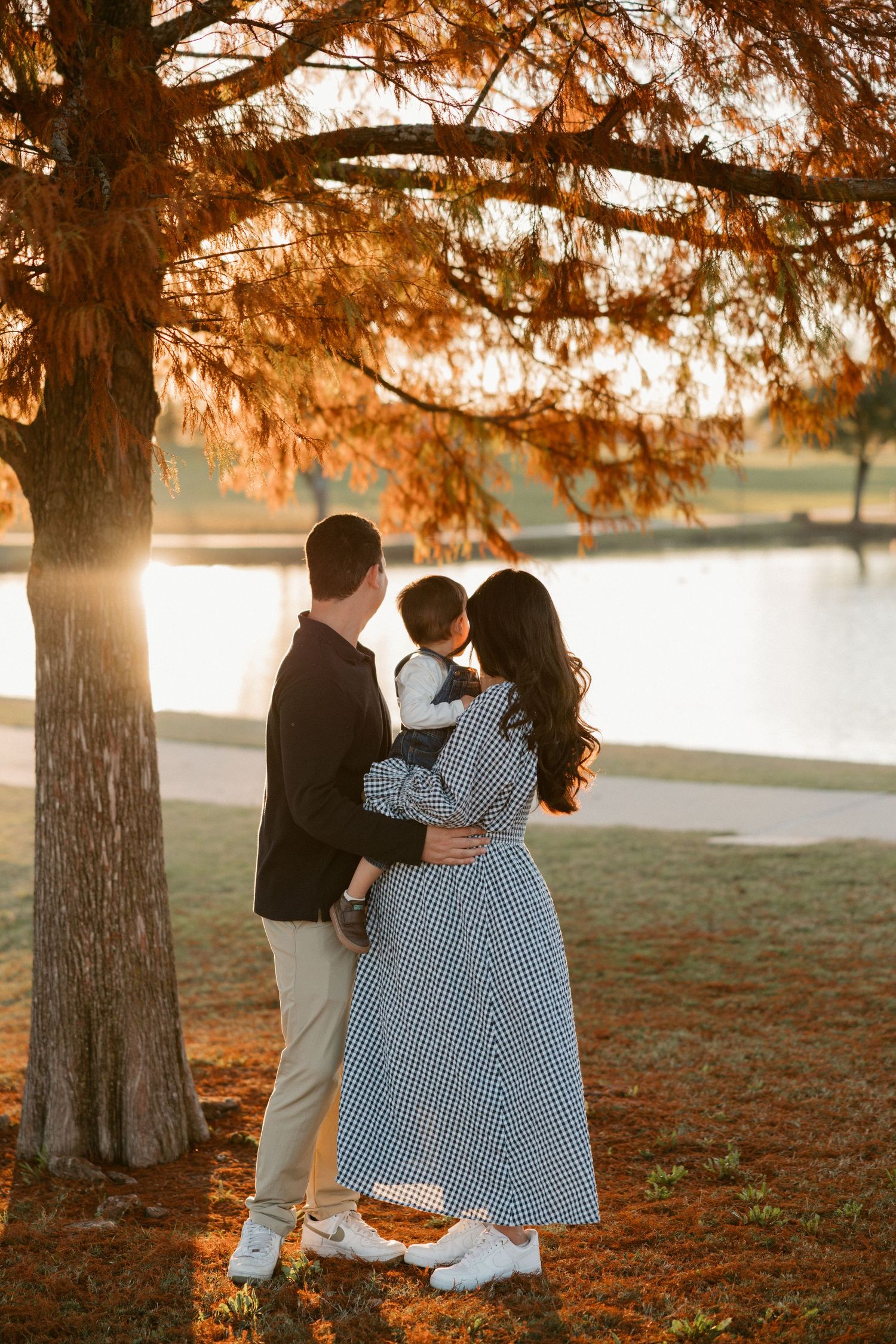 Family embraces while standing near a lake during golden autumn sunset with fallen leaves on the ground.