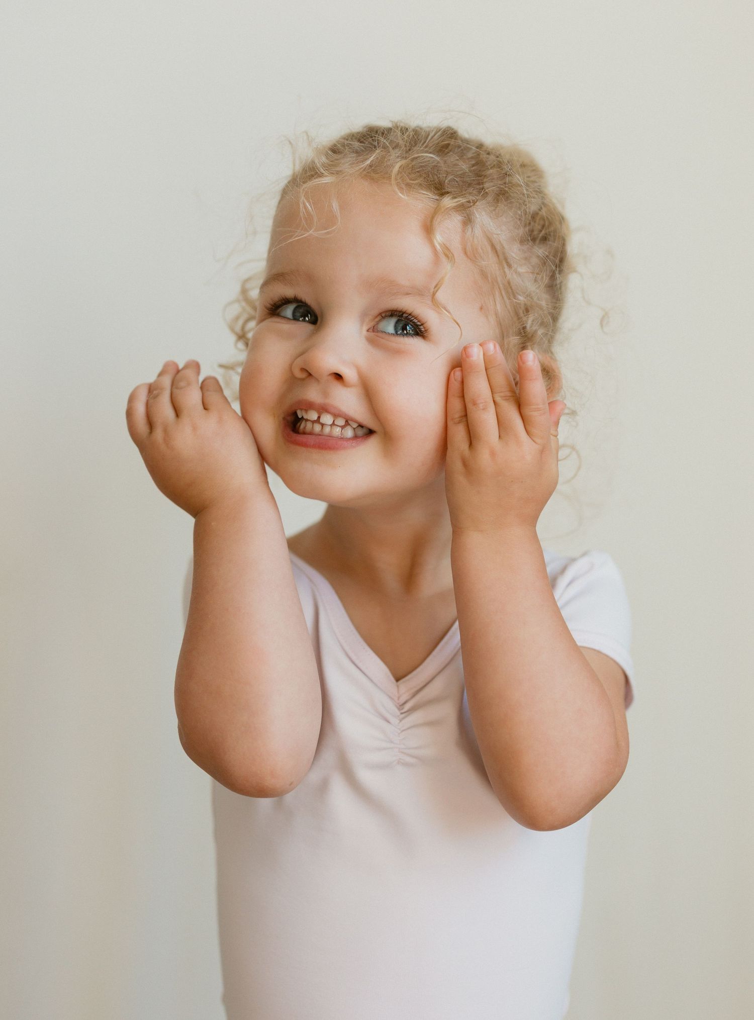 A playful child in a white shirt makes silly faces and gestures against a neutral background.