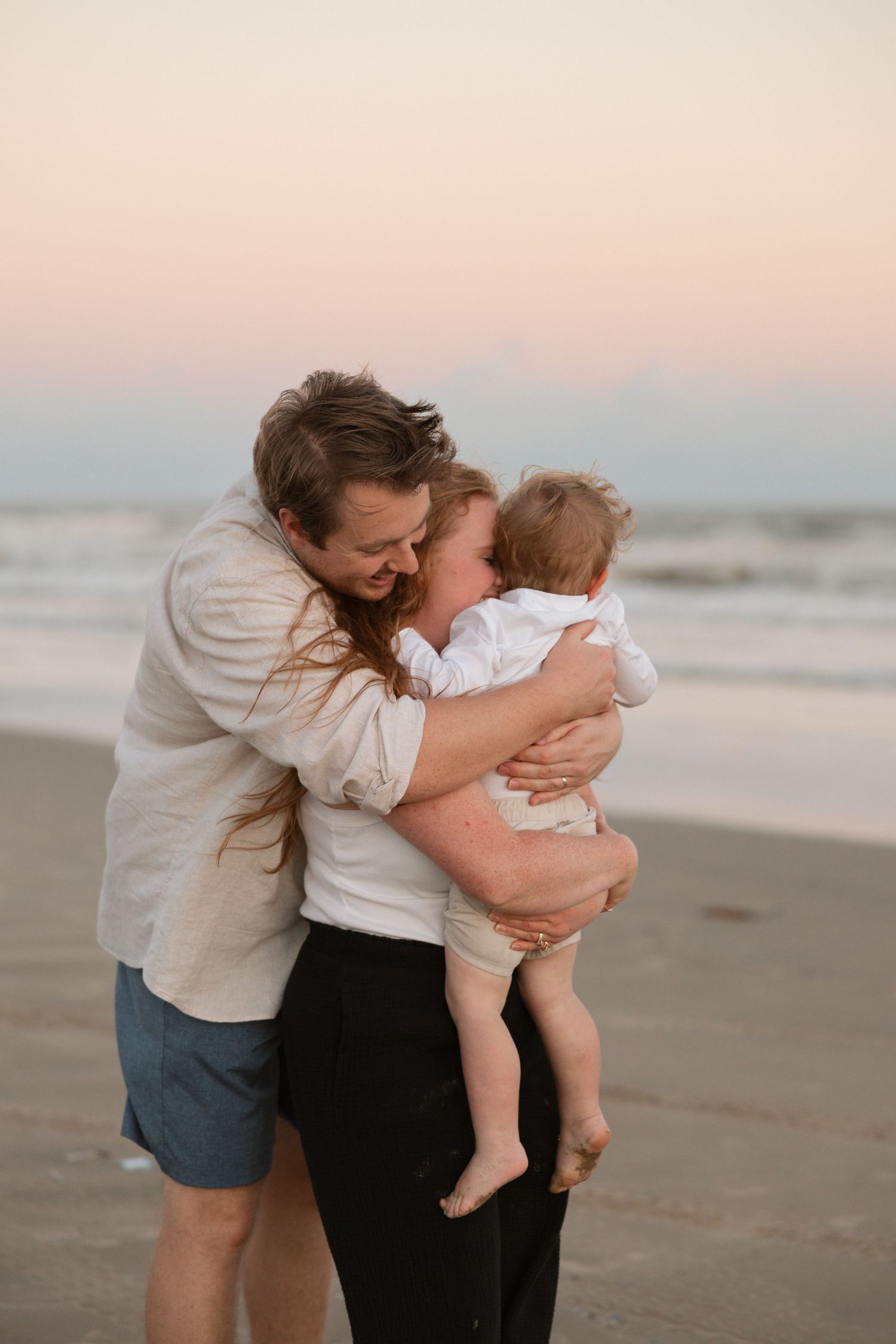 Two people share an intimate moment together on a beach at sunset with soft pink and blue sky in the background.