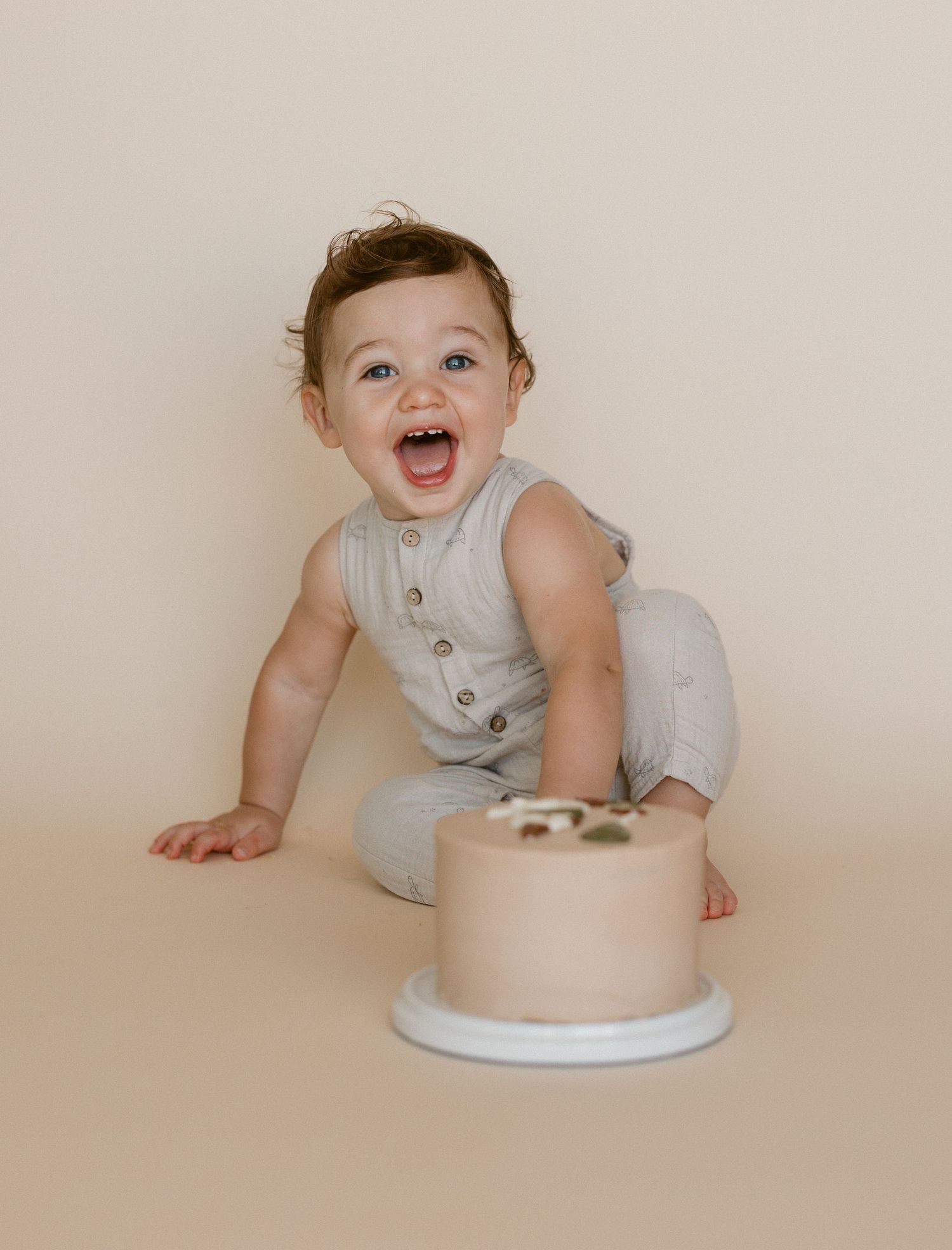 A baby in a light-colored romper crawls and plays around a simple beige cake on a white surface.
