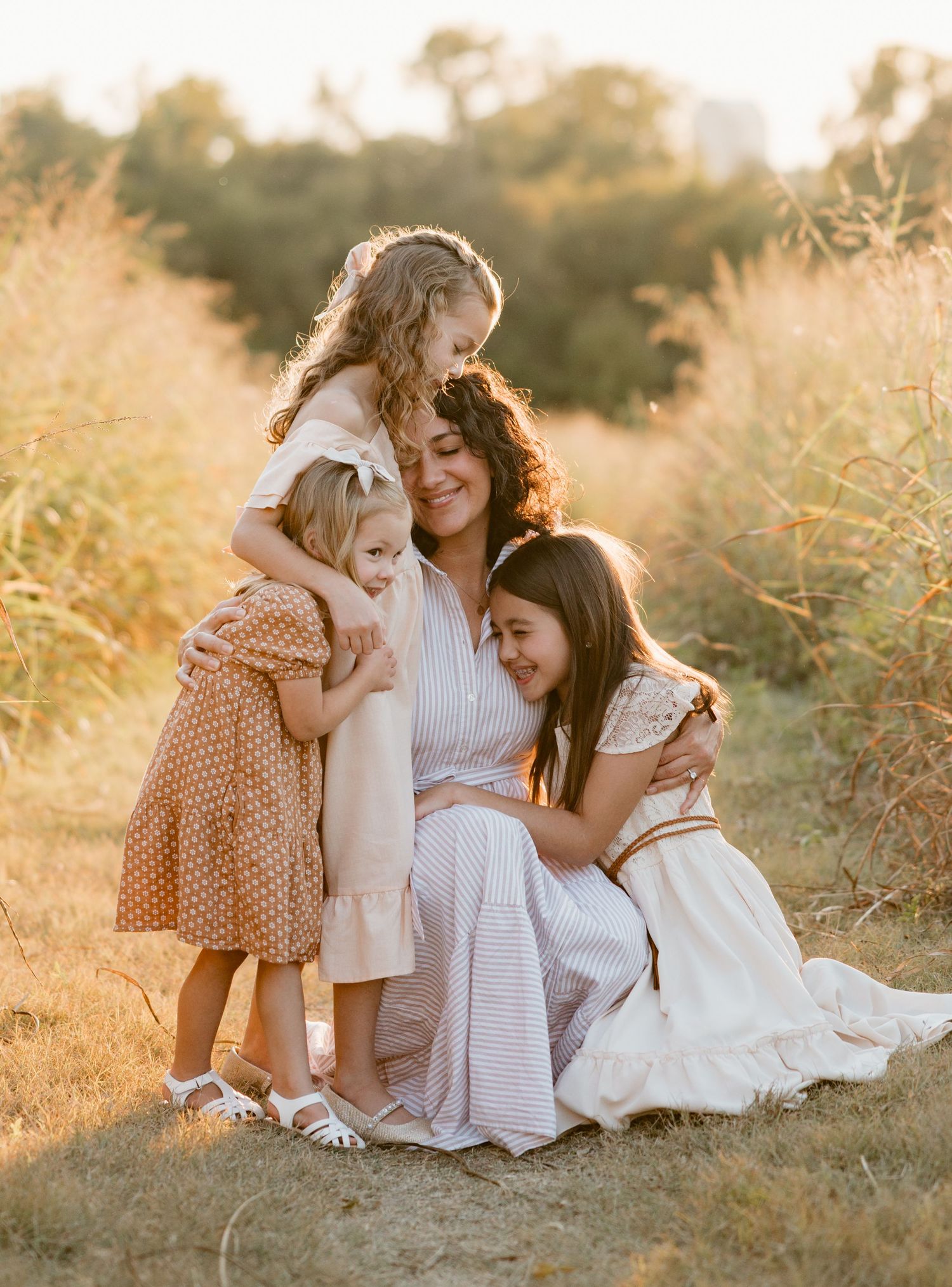 Family wearing neutral tones embraces while sitting in golden sunlit field during sunset photo session.
