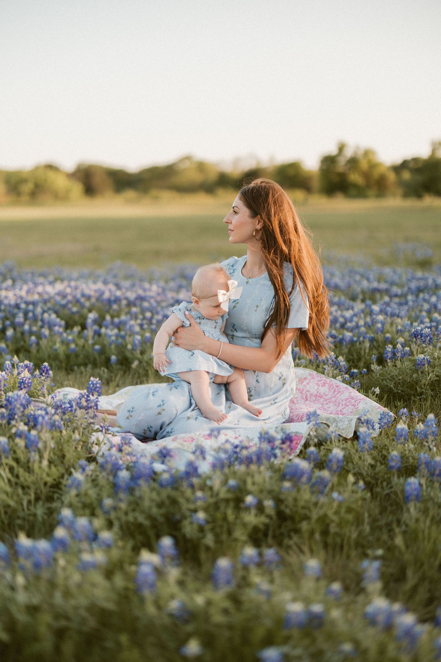 A mother with long red hair sits in a Texas bluebonnet field holding her baby, both wearing blue floral dresses at sunset.