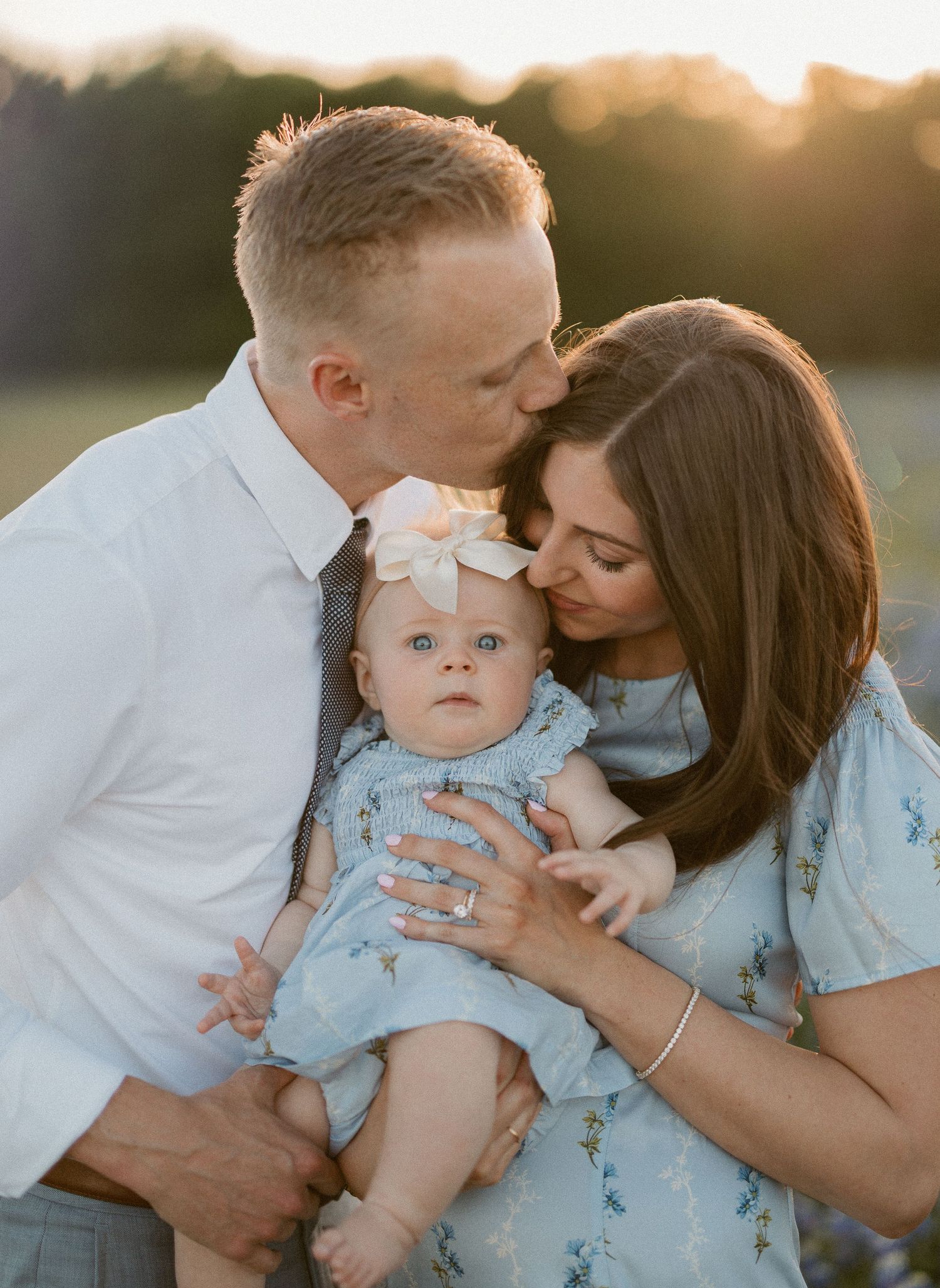 A couple kisses their baby girl in a sunlit field, mom in floral blue dress, dad in white shirt, baby wearing bow headband.