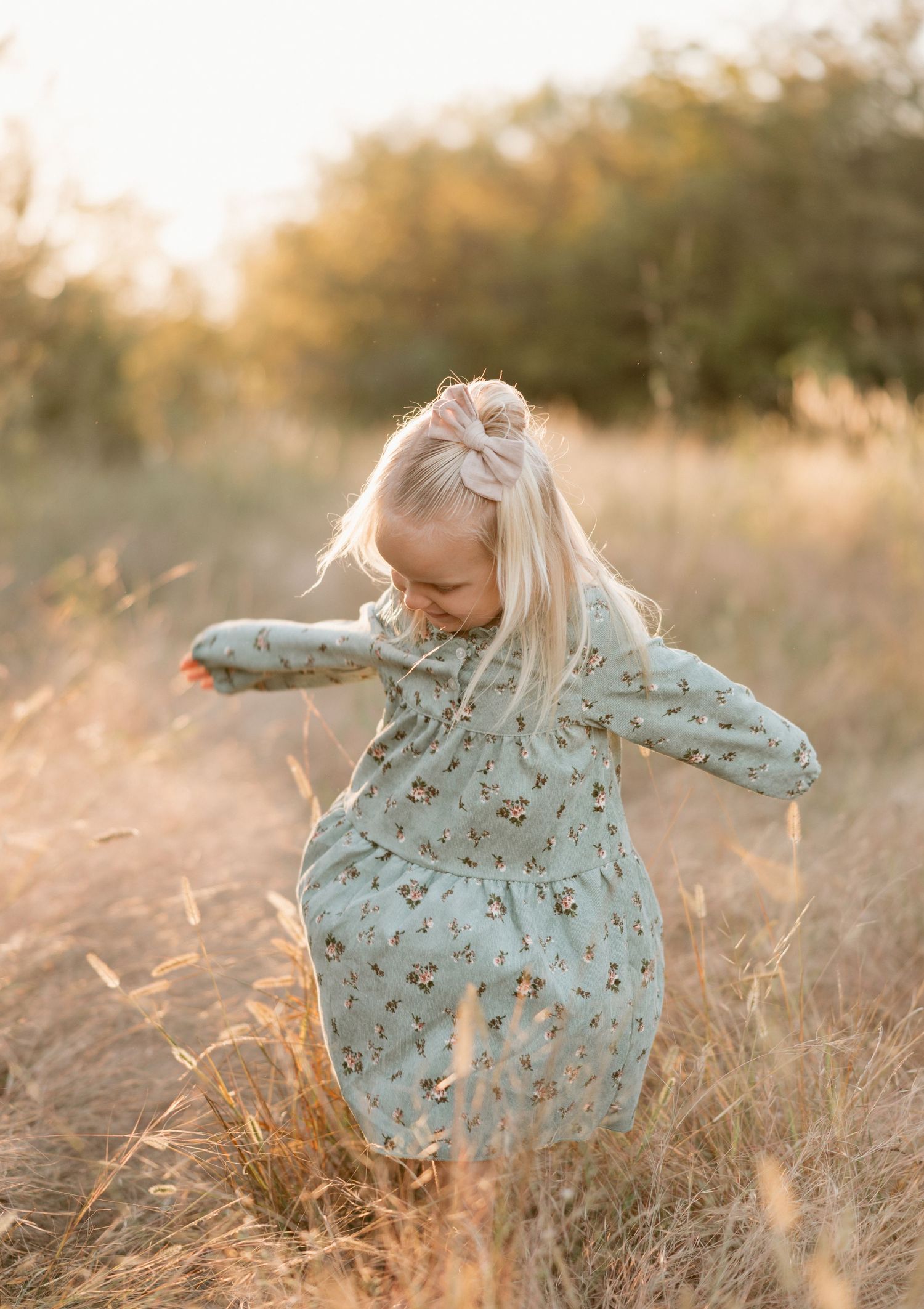 A child in a floral dress dances and twirls through a sunlit field of tall golden grass at sunset.