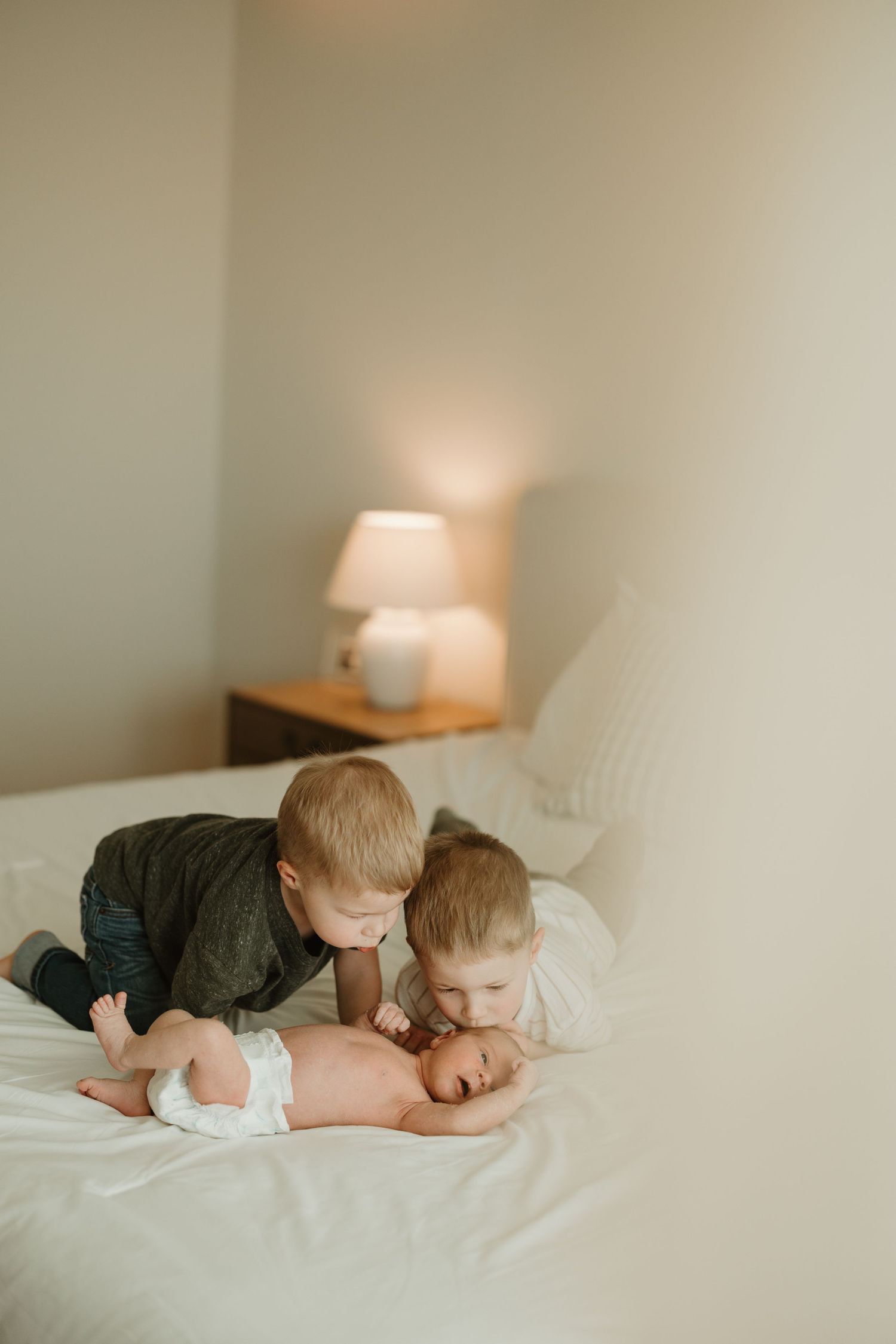 Two young children in casual clothing share a tender moment together on a white bed with a lamp in the background.