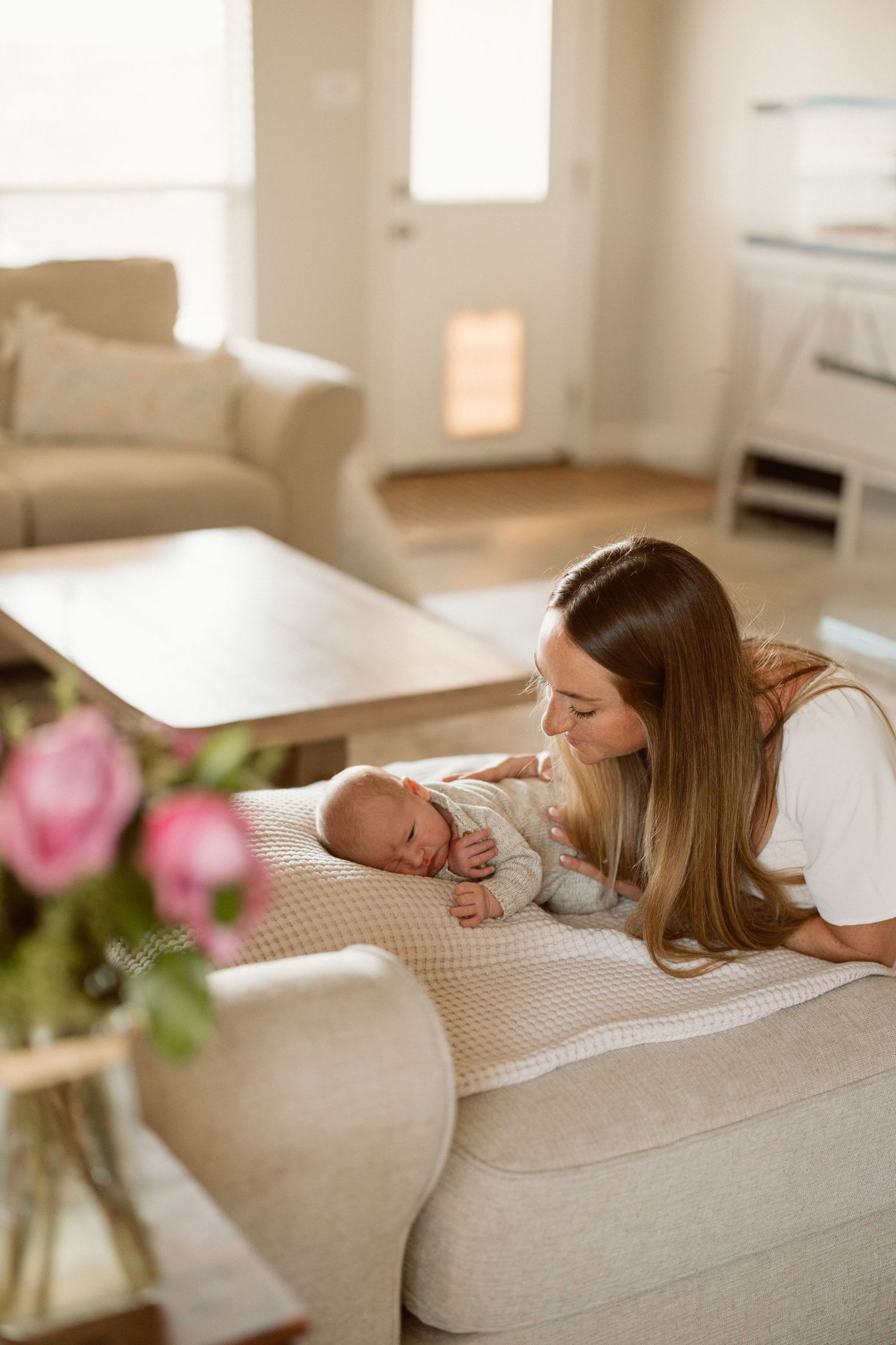 A tender moment shared between parent and newborn baby on a beige couch in a bright, cozy living room with pink flowers.