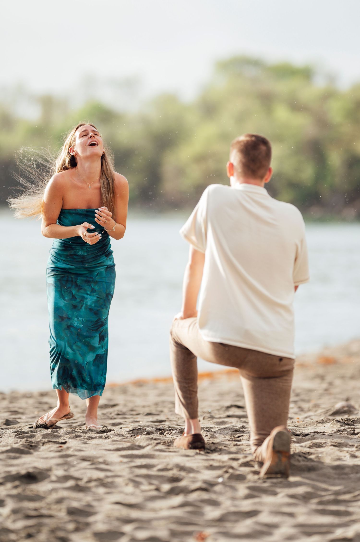 A couple shares a joyful moment during a beach proposal as they stand on sandy shores near the water.
