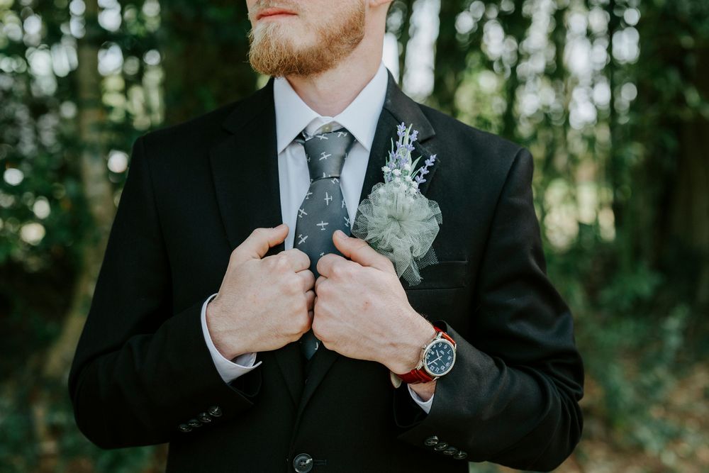 Wedding detail shots showing hands adjusting gray suit tie and boutonniere against blurred forest background.