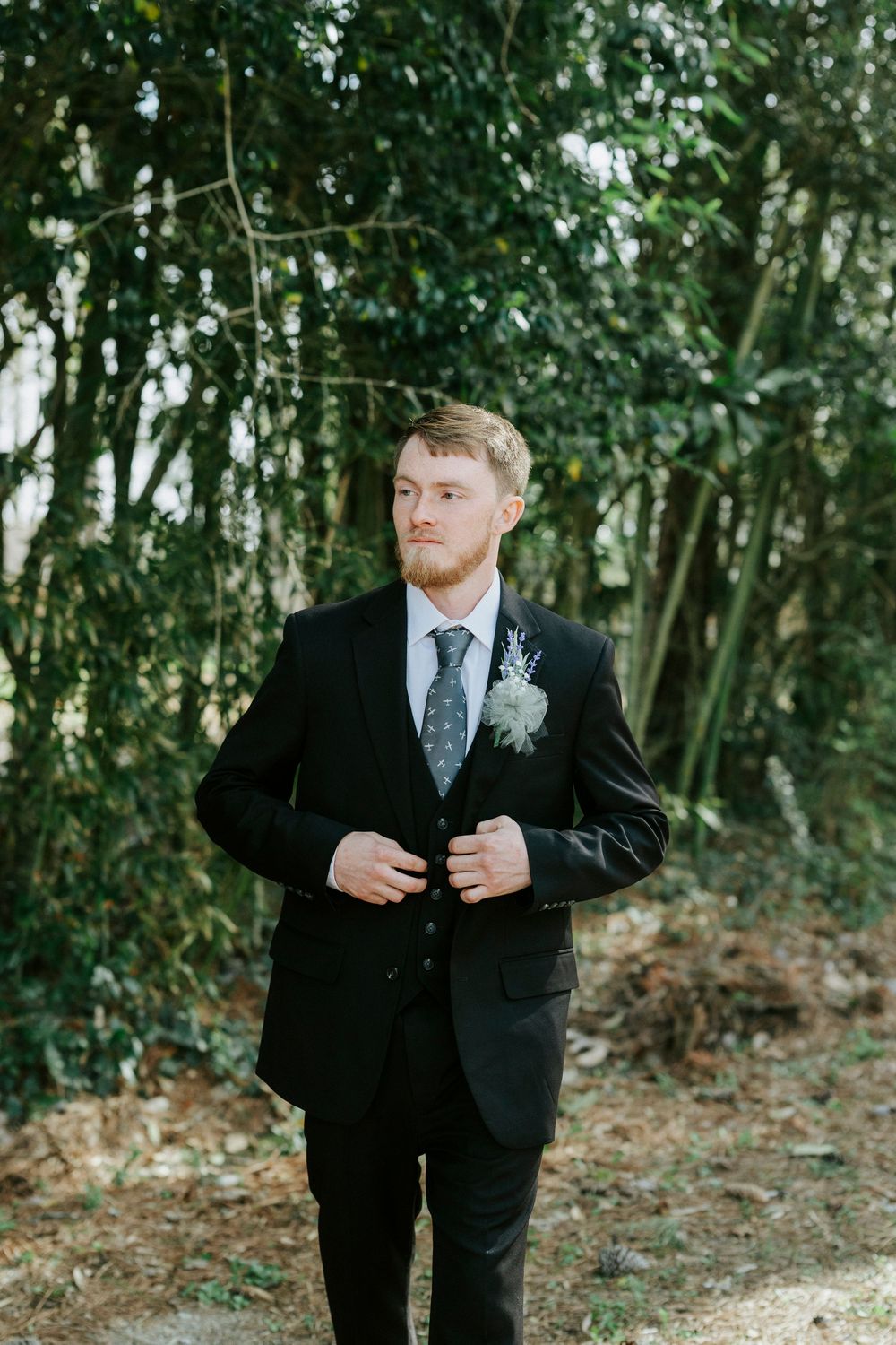 Groom in black suit and tie adjusts his jacket while standing in a wooded outdoor setting during a wedding photoshoot.