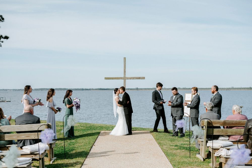 A wedding ceremony takes place outdoors by a waterfront with a white cross and wooden benches on a grassy lawn.