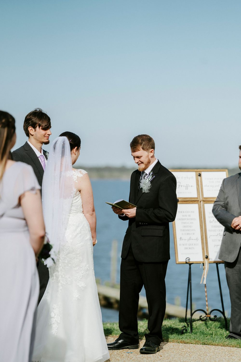 An outdoor waterfront wedding ceremony with a couple and officiant standing near a decorative easel sign.