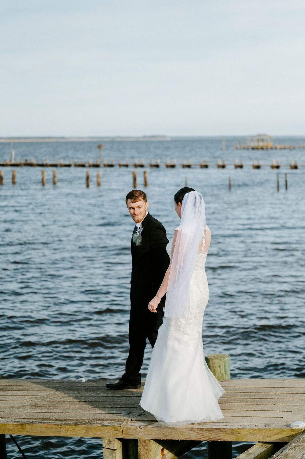 A couple sits on a wooden dock overlooking the ocean during a wedding photoshoot at sunset.