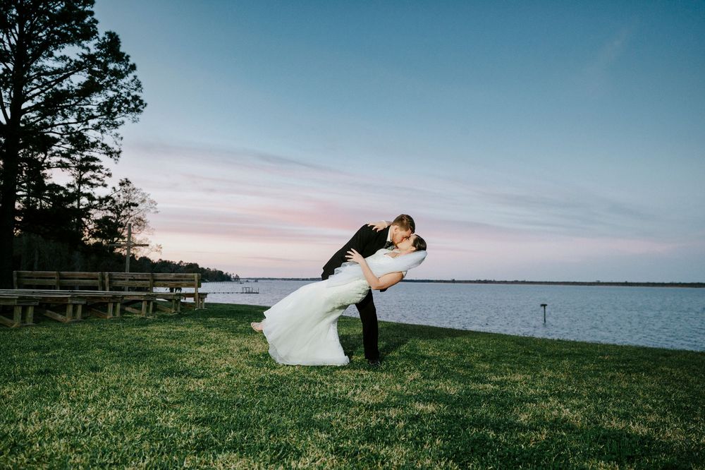 A romantic wedding couple embraces on a grassy hillside overlooking a serene lake at sunset.