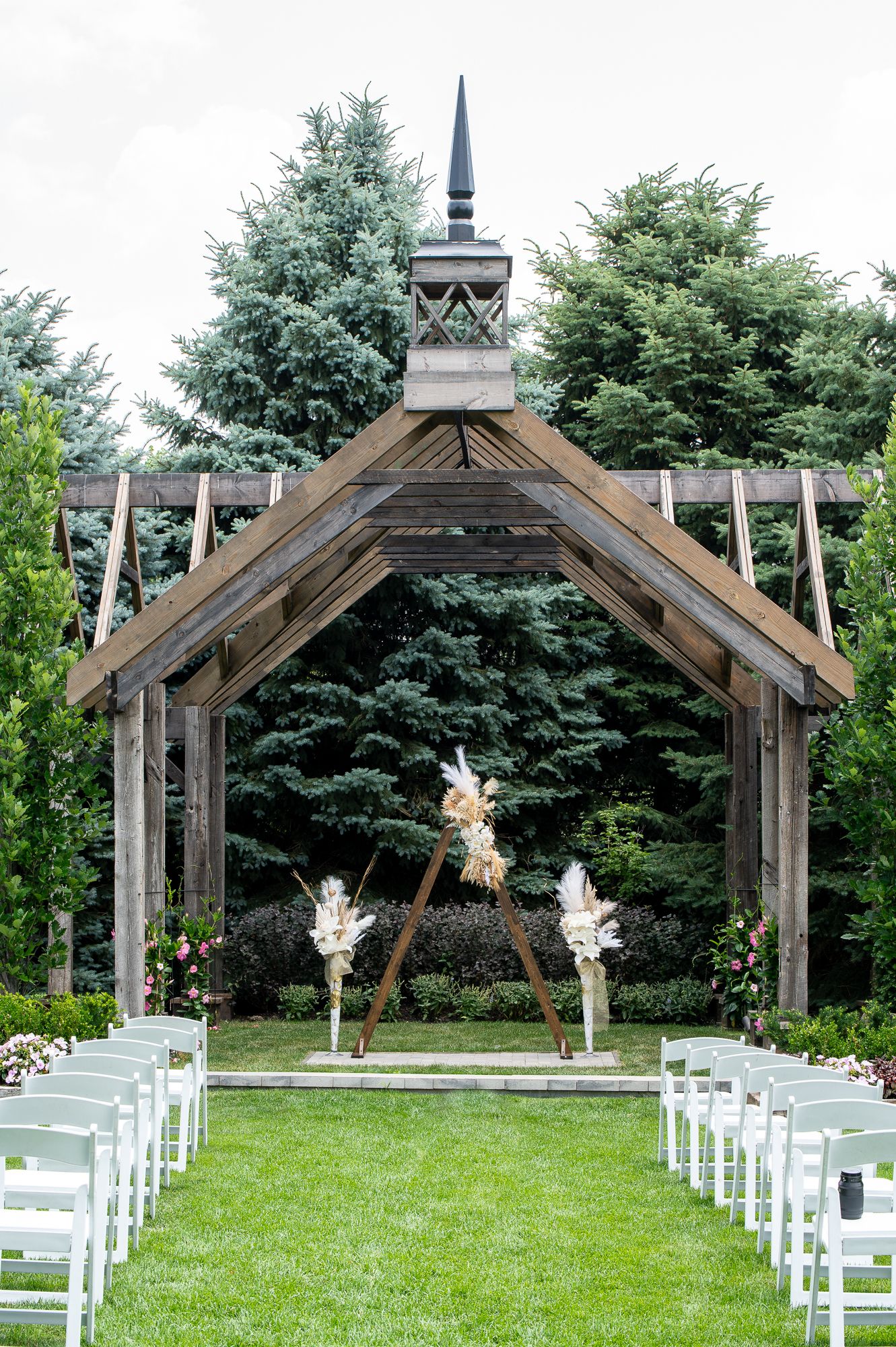 Rustic wooden wedding ceremony pavilion with triangular floral arch, white chairs on green lawn, surrounded by lush evergreen trees.