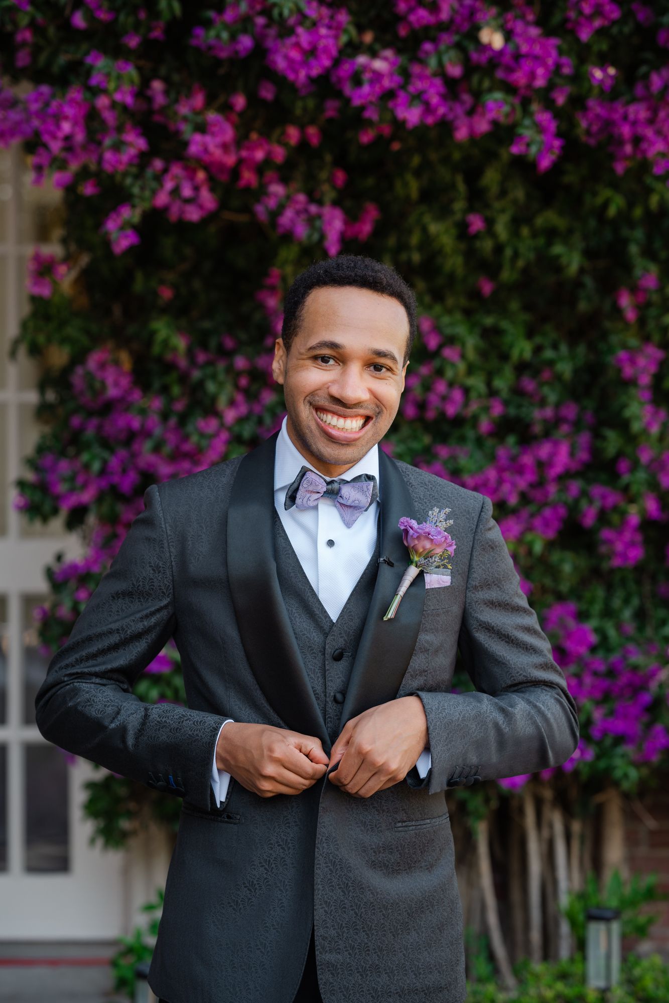 A groom in a charcoal grey suit and purple bowtie adjusts his collar in an elegant wood-paneled room.