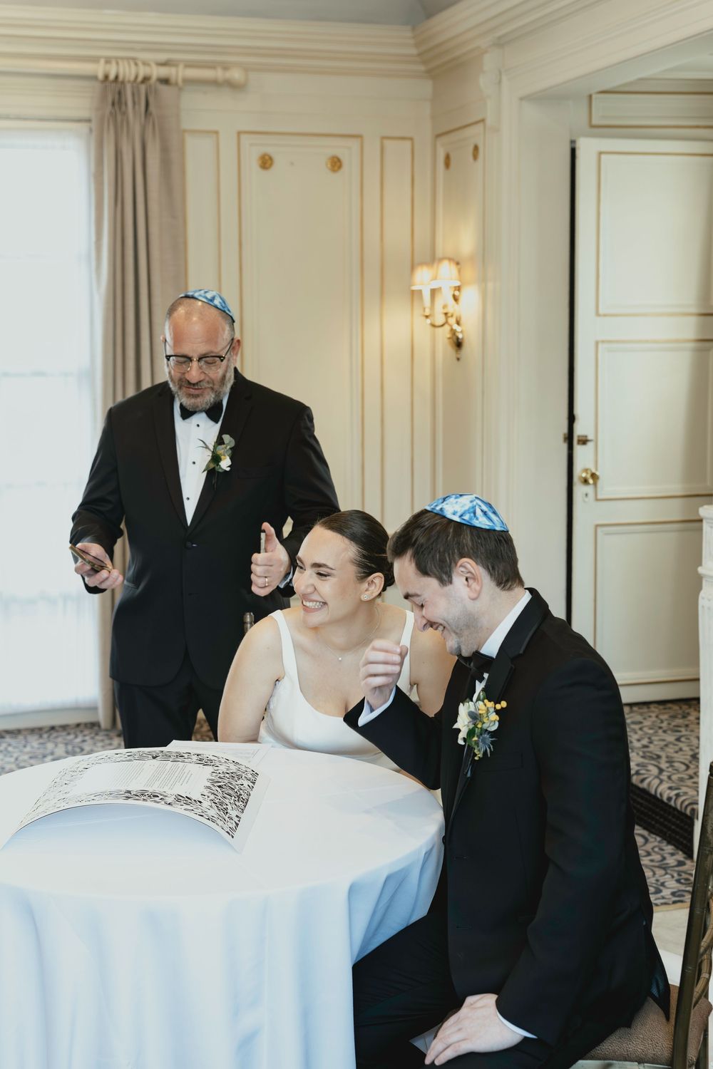 Wedding party members in formal black attire share a candid laughing moment at a reception with white tablecloths.