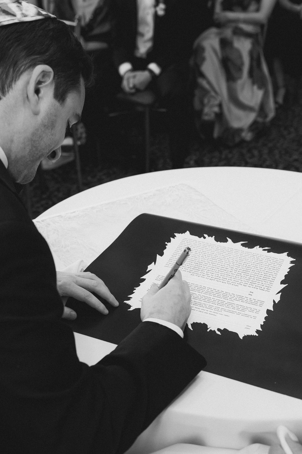 A person in formal attire signs documents on a white surface against a dark background.