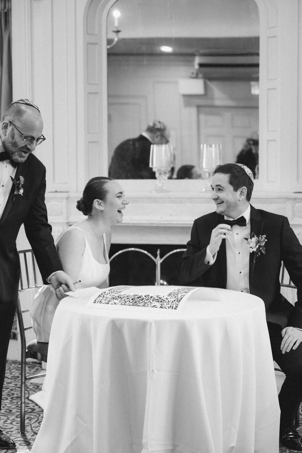 A black and white photograph shows people laughing together at round tables covered with white tablecloths.