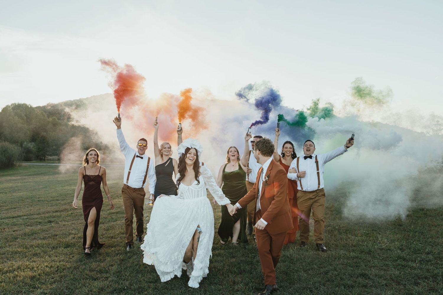 Wedding party walks through colorful smoke bombs in a grassy field at sunset creating a dreamy atmosphere.