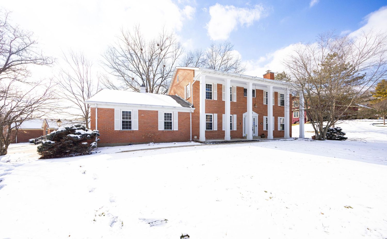 A panoramic winter landscape shows a snow-covered field with brick buildings and bare trees in the background.