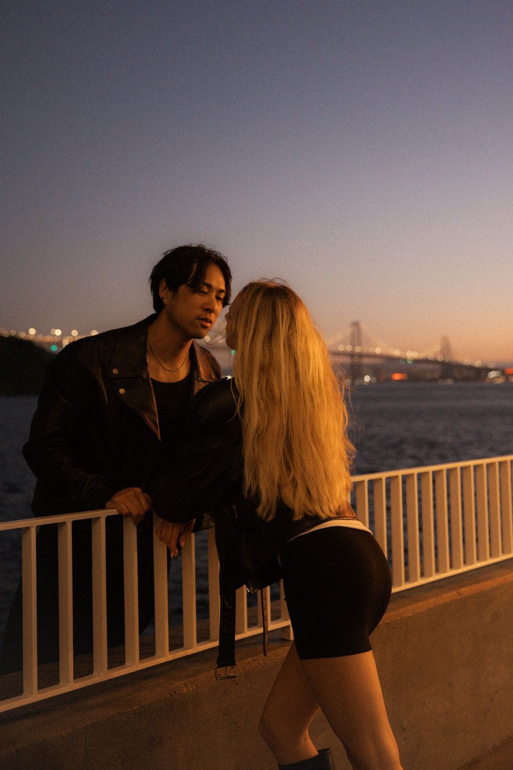 Couple standing by waterfront railing at dusk, woman in black mini dress, man in leather jacket, city lights behind.