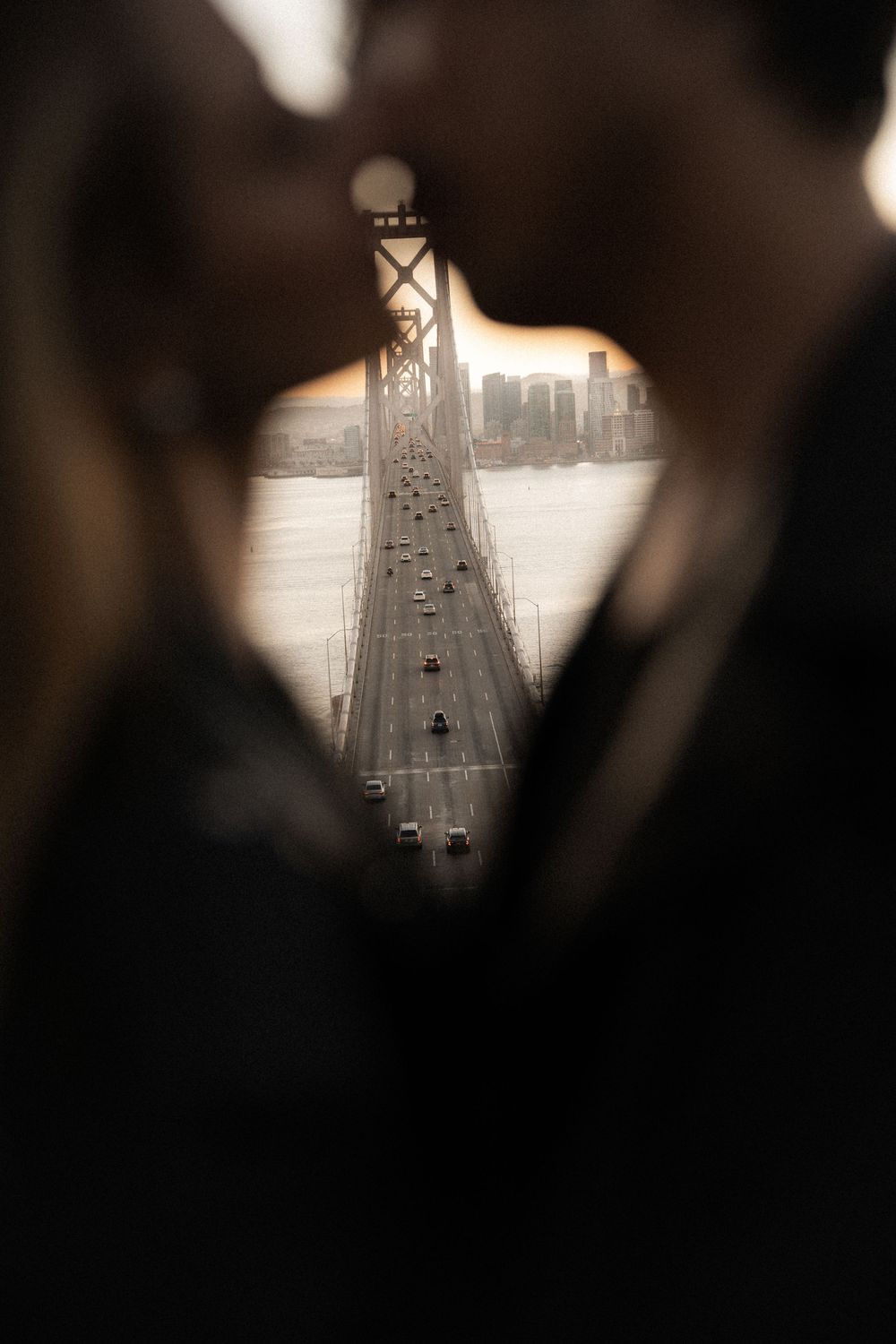 Romantic couple kissing with San Francisco Bay Bridge traffic and city skyline framed between them at golden hour.