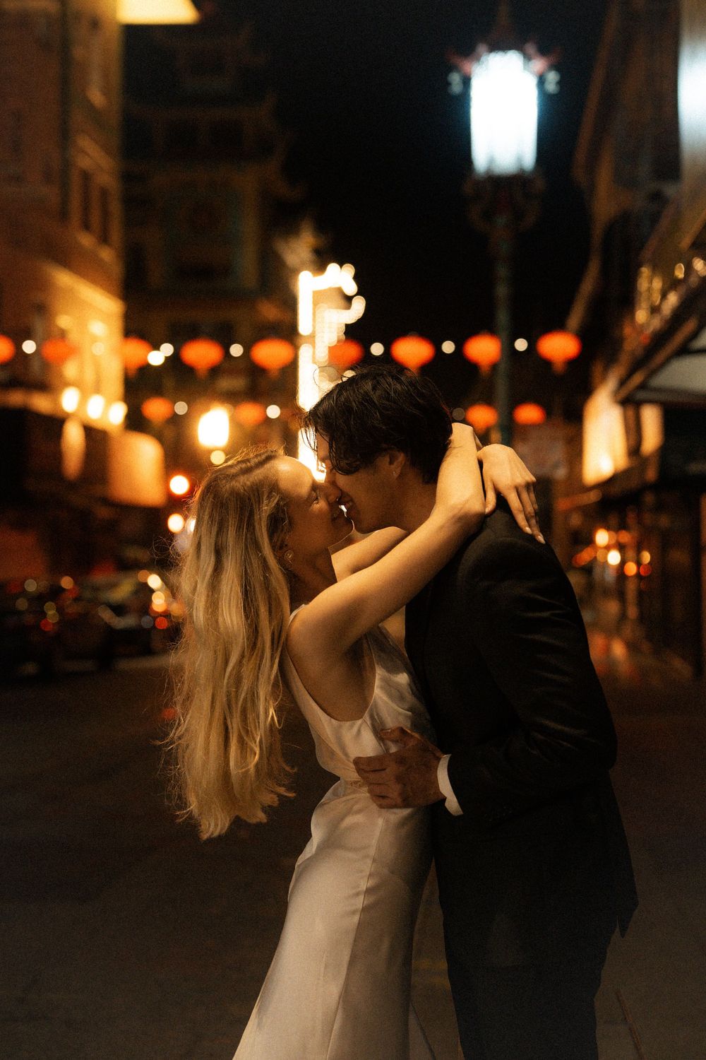Couple sharing a romantic kiss at night with glowing red lanterns illuminating a city street behind them.