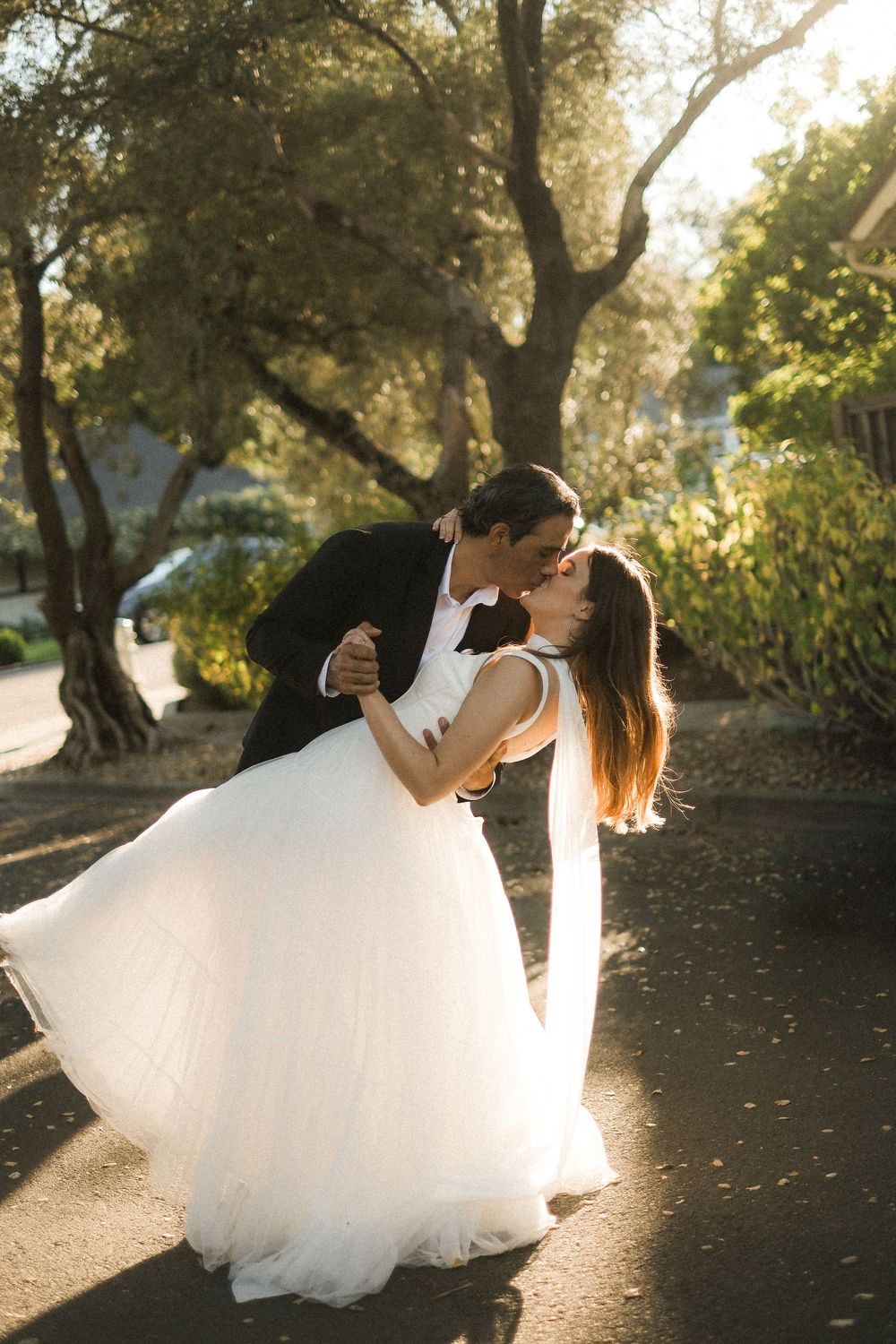 Romantic wedding couple dips and embraces in golden sunset light under trees.