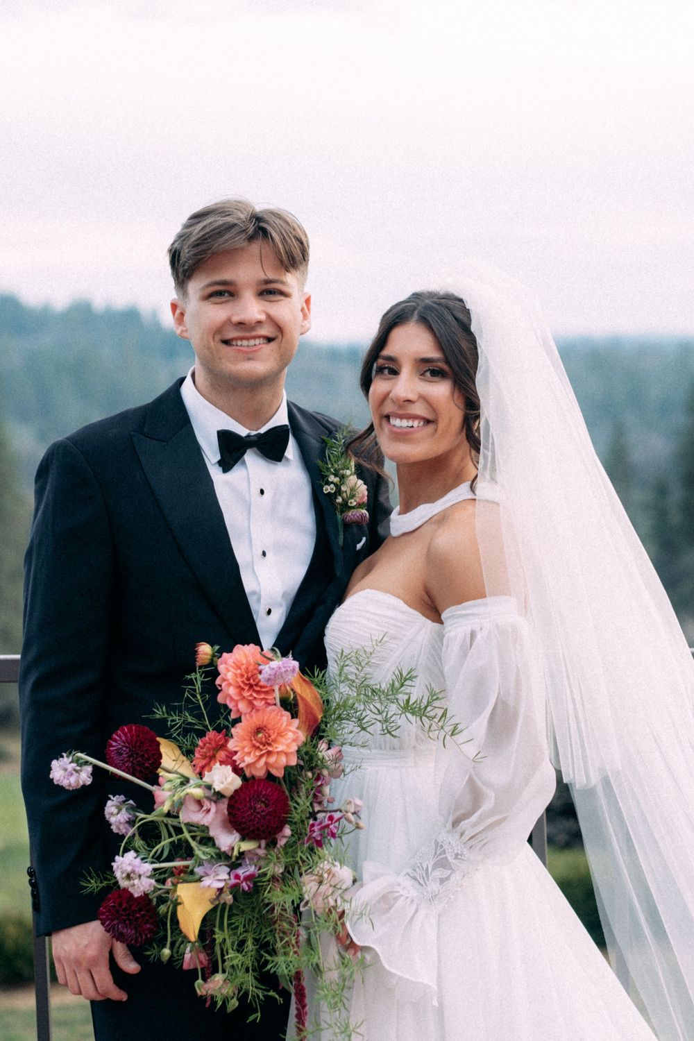 Newlyweds pose together outdoors holding vibrant floral bouquet against mountain backdrop.