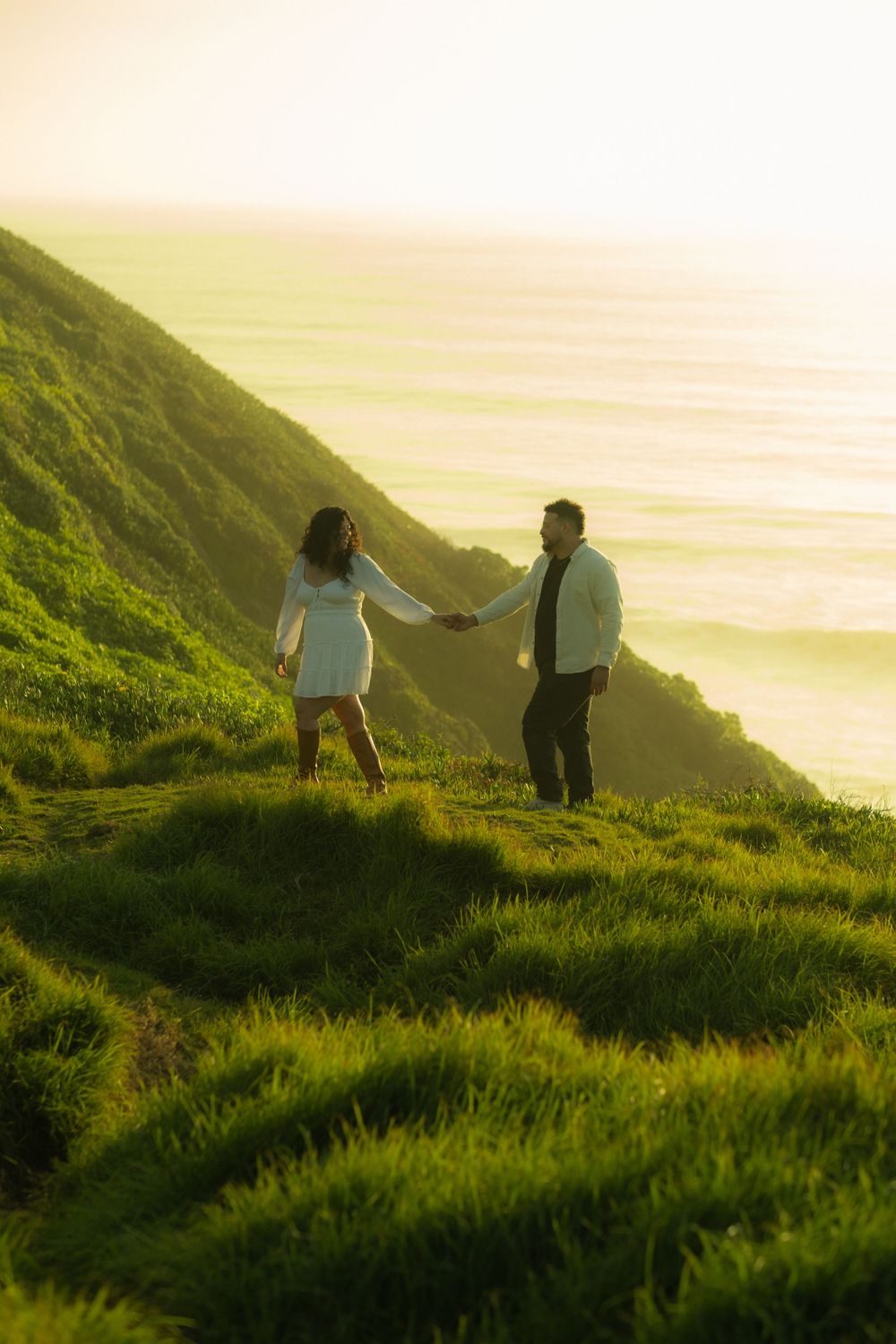 Two people stand on a grassy hilltop overlooking the ocean at sunset.