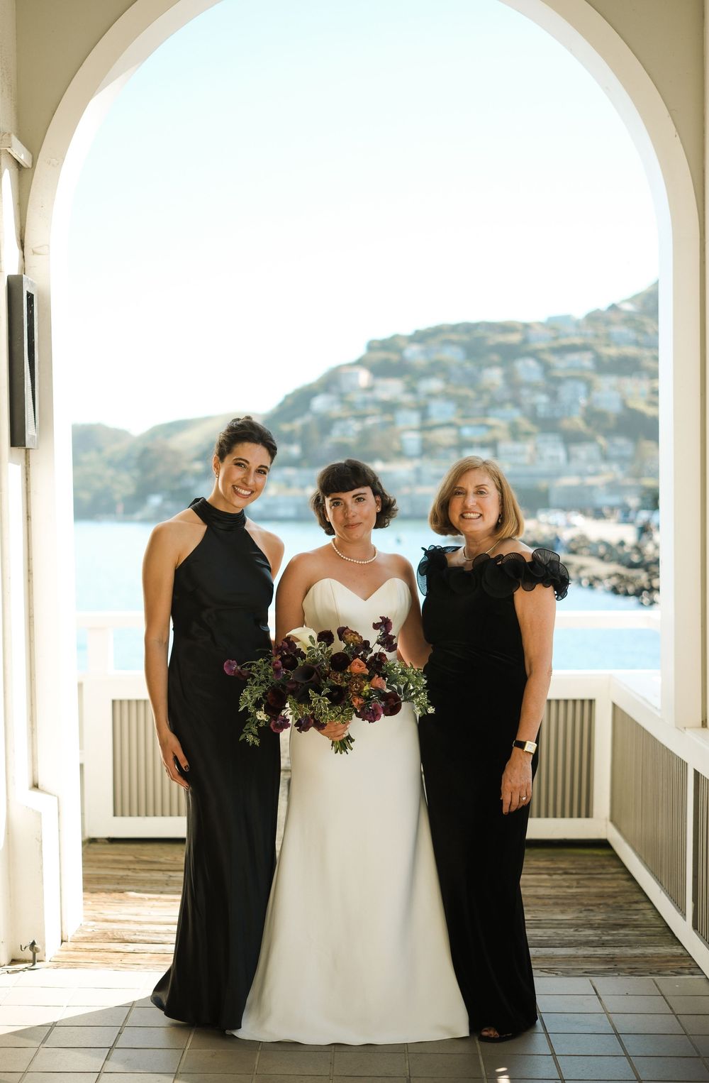 Bride in white gown poses with two women in black dresses, holding bouquet, with coastal view backdrop.