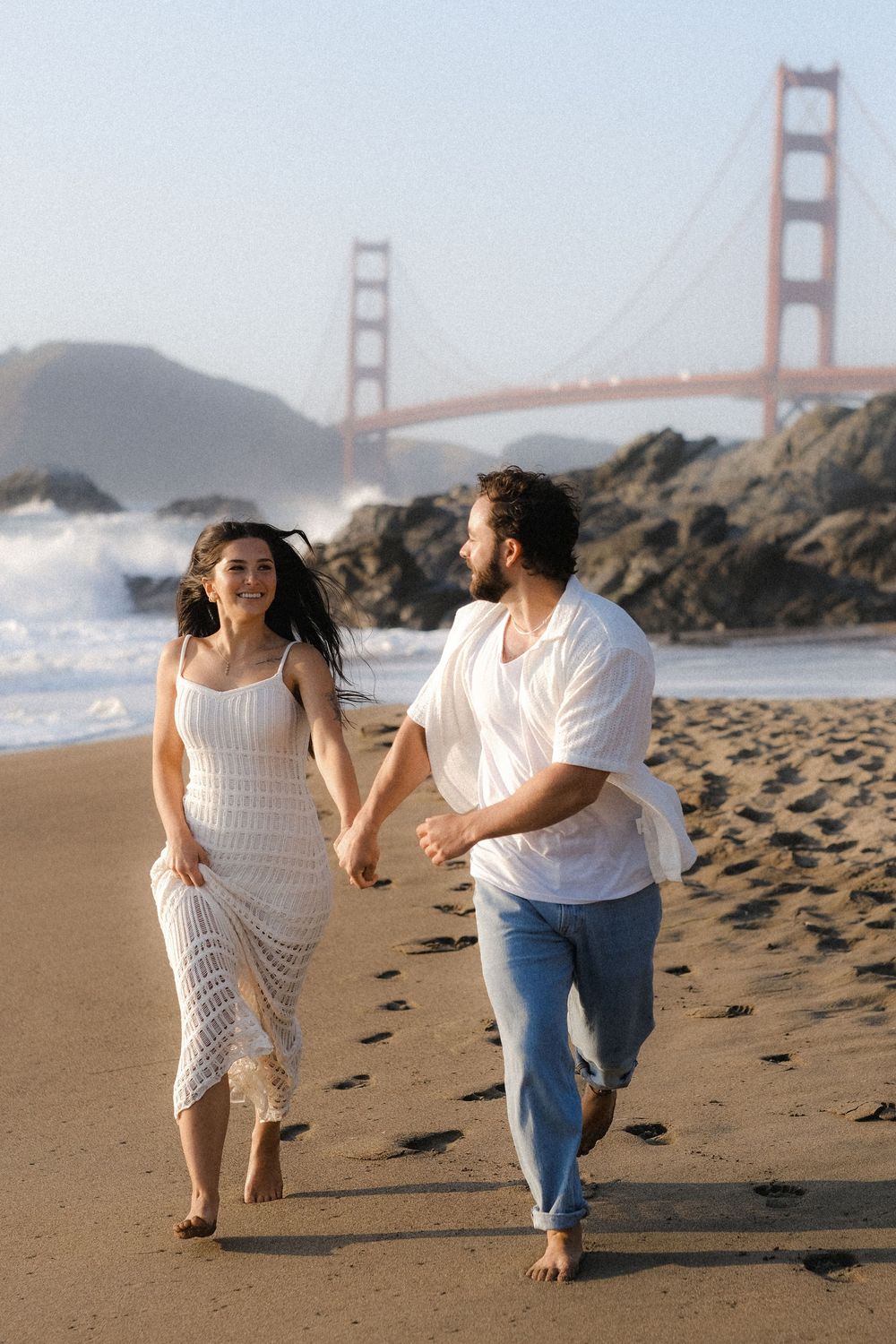 A couple runs playfully along Baker Beach with the Golden Gate Bridge visible in the foggy San Francisco background.