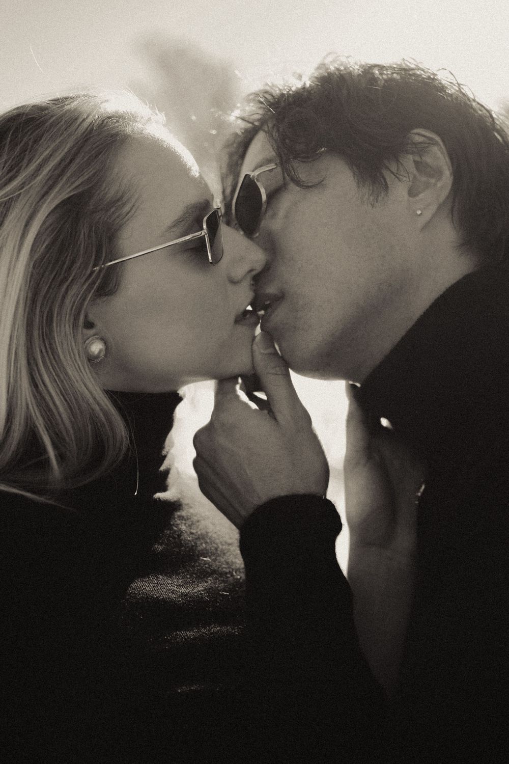 Black and white photo of a couple sharing a cigarette kiss, both wearing sunglasses in a romantic outdoor setting.