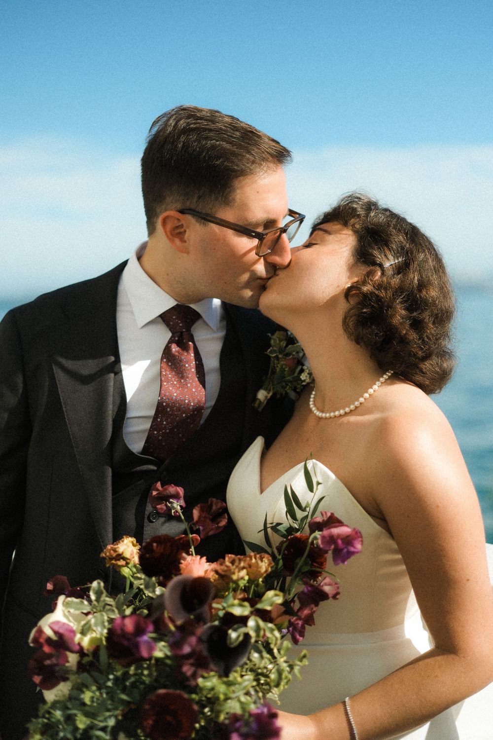 Bride and groom sharing a kiss outdoors, bride holding a lush burgundy bouquet, blue sky and water in the background.