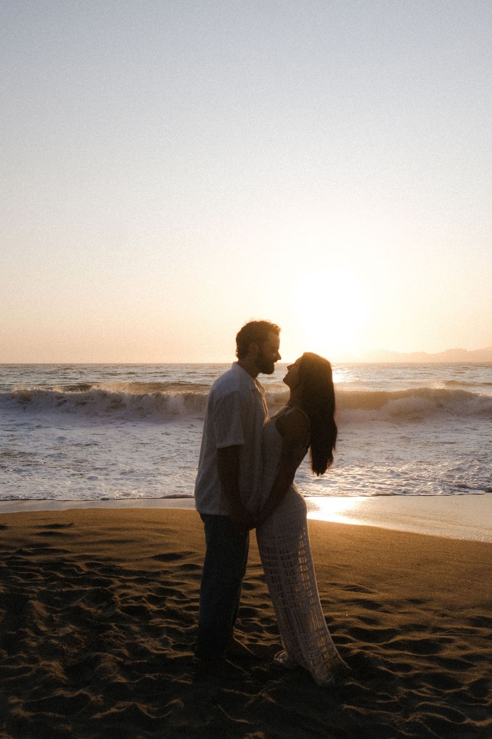 A silhouetted couple embraces on a sandy beach at sunset with ocean waves crashing in the background.